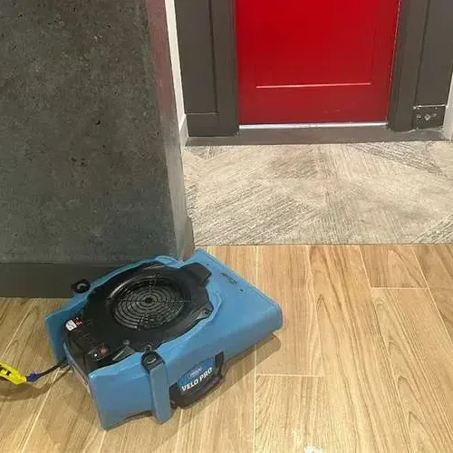 A blue air mover running on a wood-look tile floor beside an open doorway, drying the room after a water loss